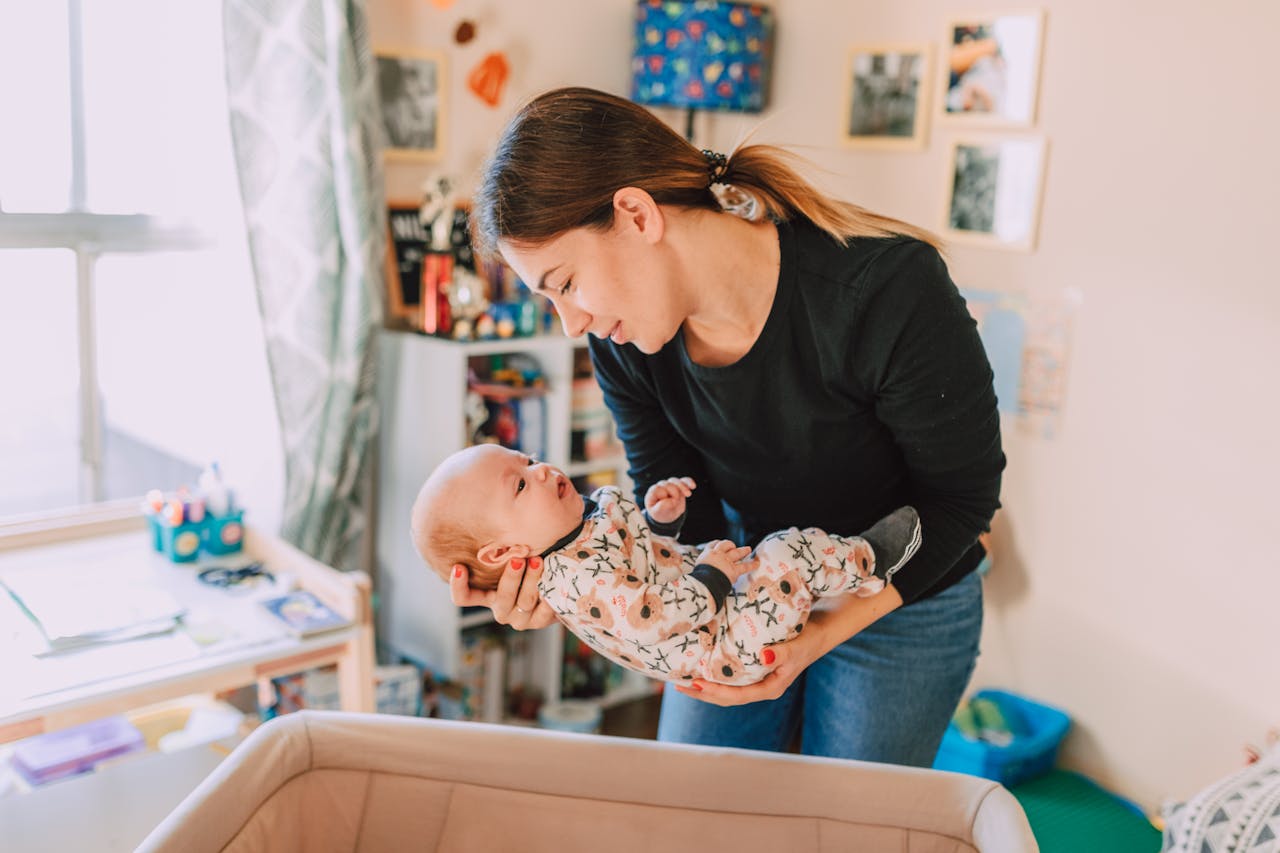 Une mère tient son bébé dans les bras au‑dessus du berceau, dans une chambre d’enfant lumineuse, en le regardant avec un sourire tendre